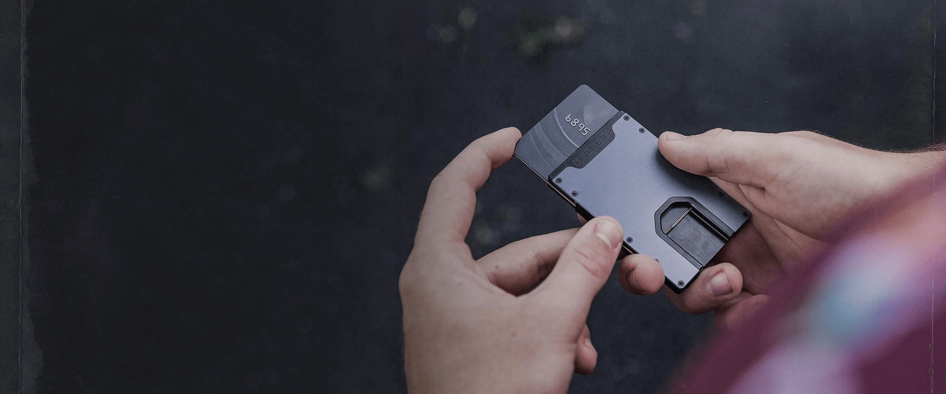 A man’s hands hold a steel gray GeoGrit wallet with a dark gray credit card sliding out of the top.